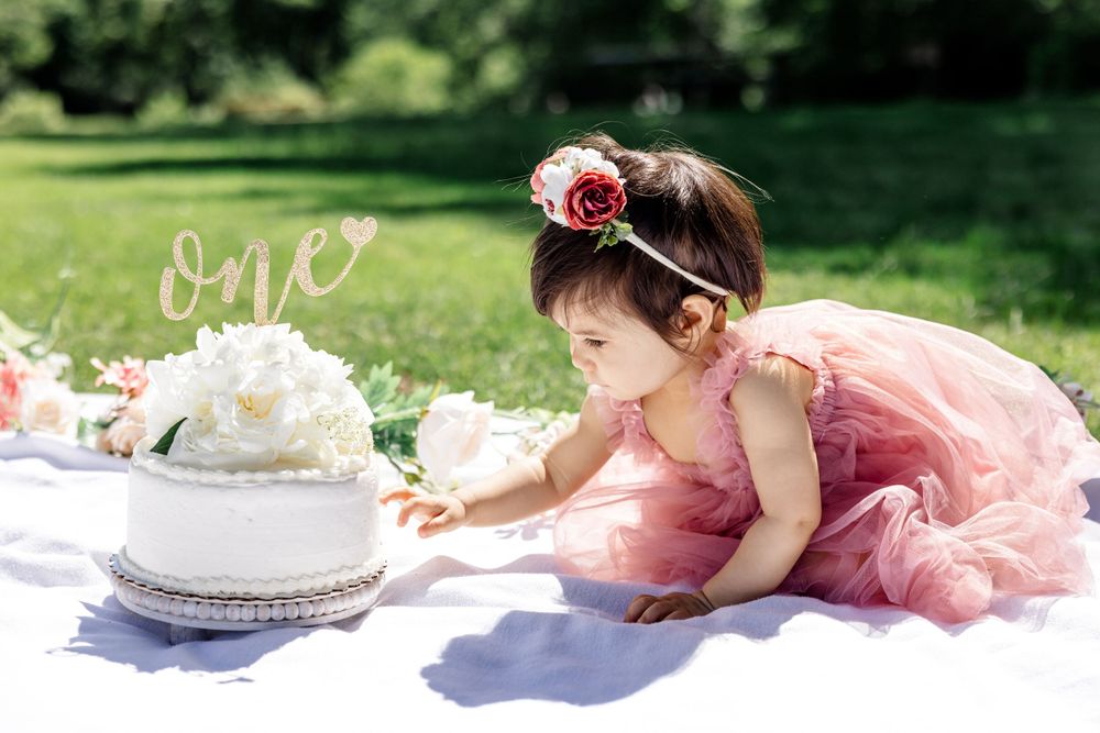 Young child in pink tulle dress reaches toward white birthday cake on grass.