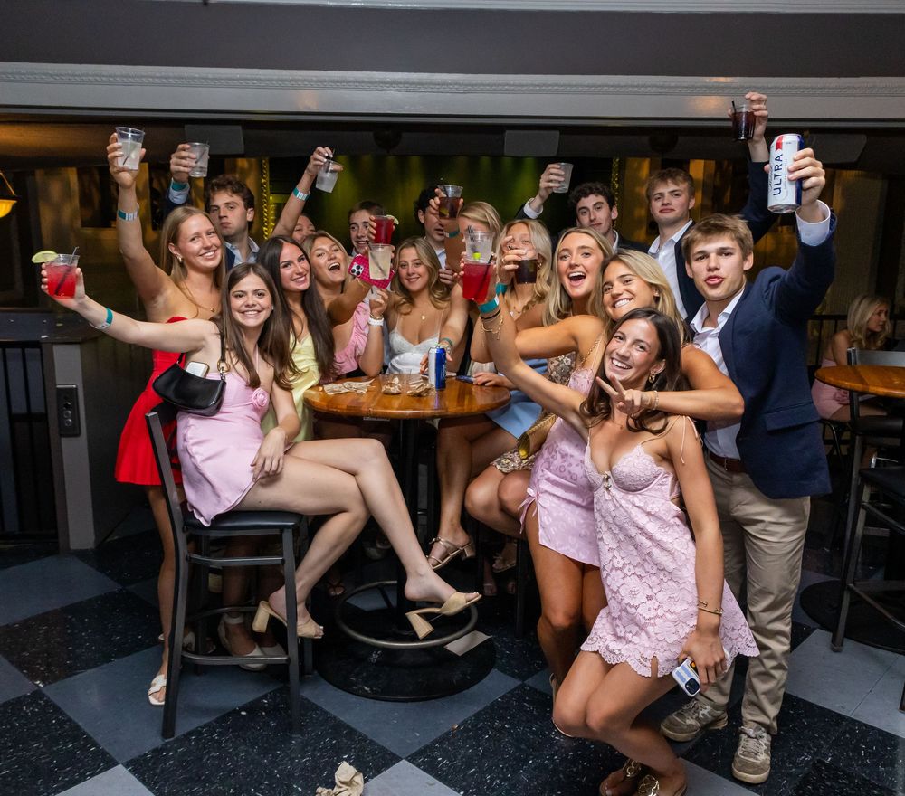 Large group of young adults celebrating at a bar, raising colorful drinks and smiling together.