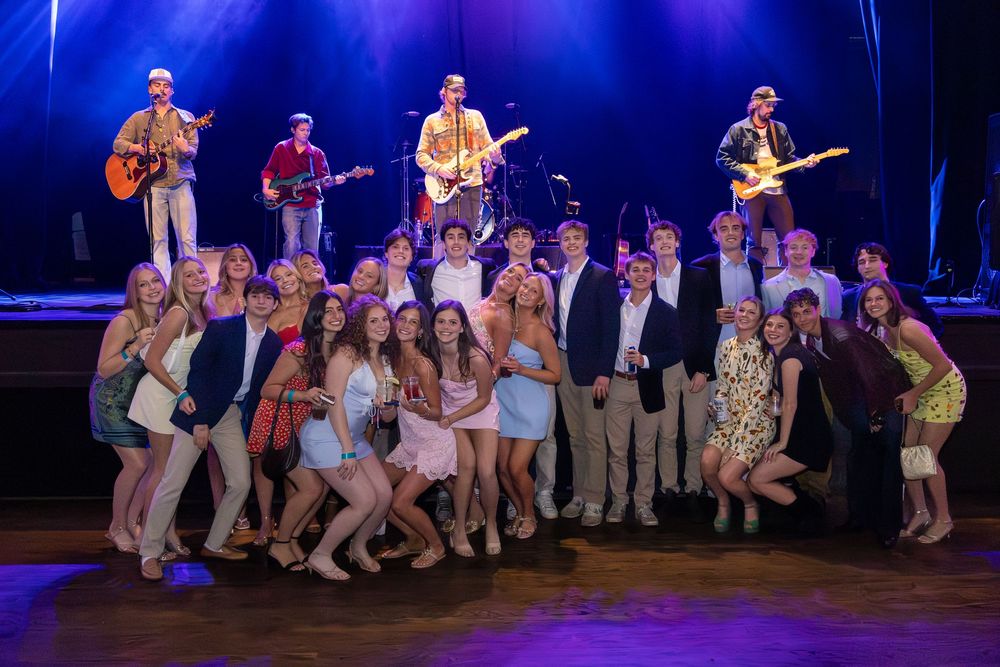 Large group of teens posing on stage with a live band performing behind them at a formal event.