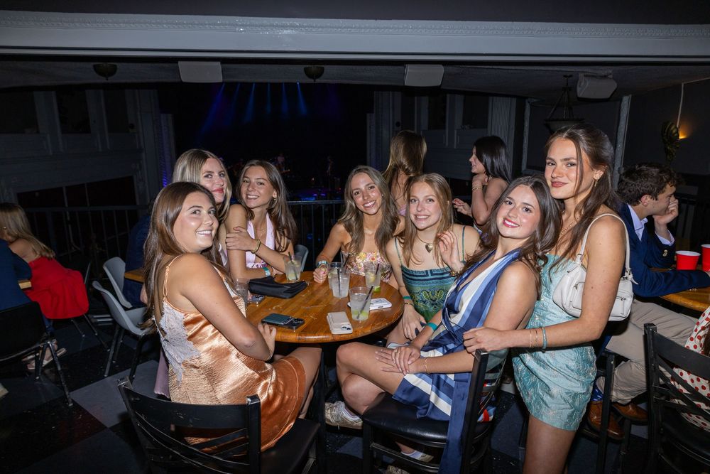 Group of young women smiling and posing together at a table during a nighttime social gathering.