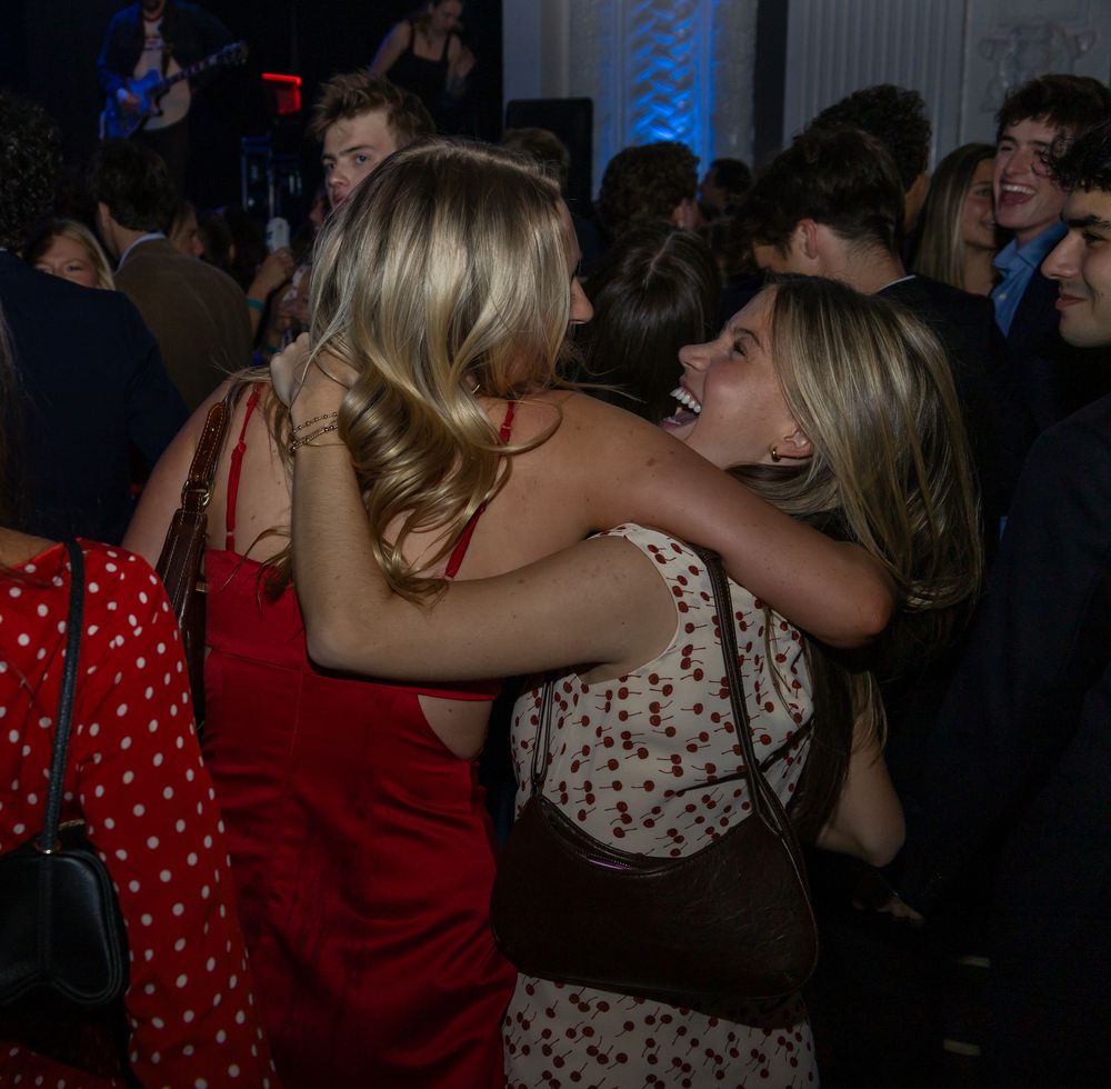 Two blonde women hugging and laughing at a party, wearing red and floral dresses in a dimly lit venue.