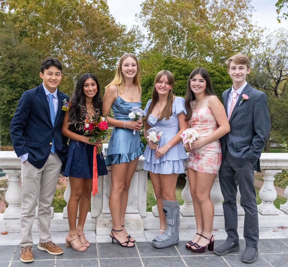 Group of six teenagers dressed up for homecoming posing together outdoors holding floral bouquets.