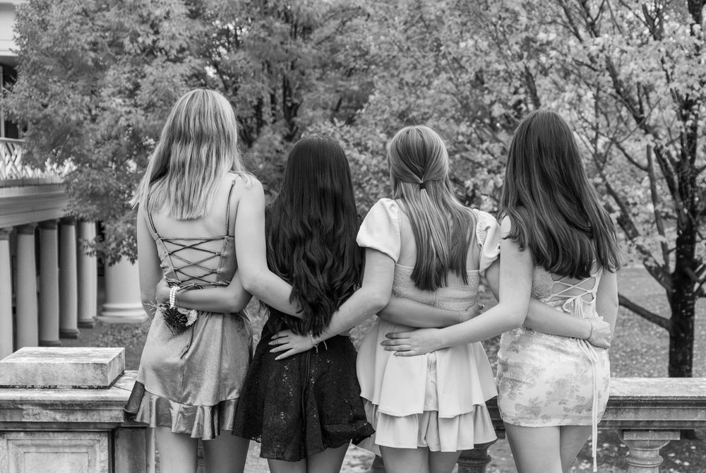 Four girls in dresses stand arm in arm with their backs to the camera in a black and white photo.