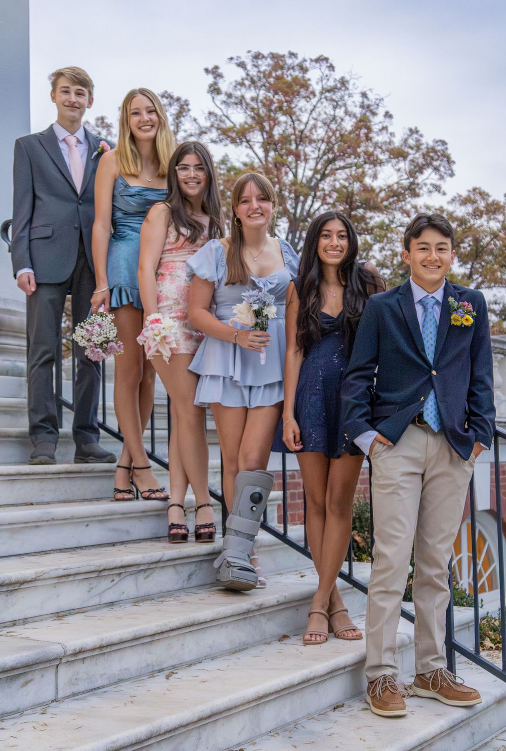 Six teenagers dressed in formal attire pose on stone steps for a prom or homecoming photo.
