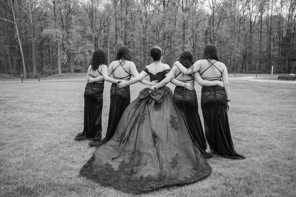 Five women in formal gowns pose with backs to camera in a black and white outdoor wedding photo.