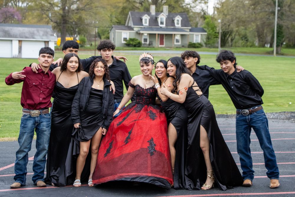 A quinceañera girl in a red and black ball gown poses with her court of honor on a track field.