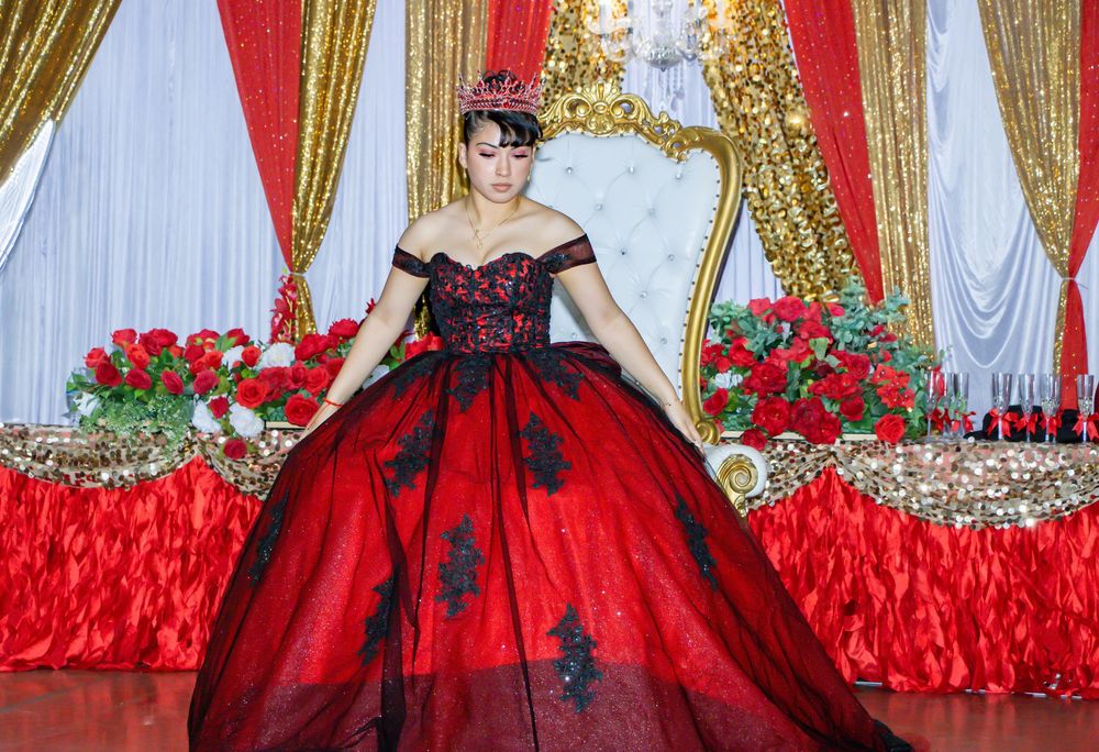 A quinceañera girl in a red and black ball gown poses regally on a throne at her reception venue.