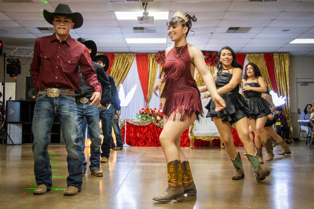 People line dancing in cowboy boots and western attire at a festive indoor celebration with red and gold decorations.