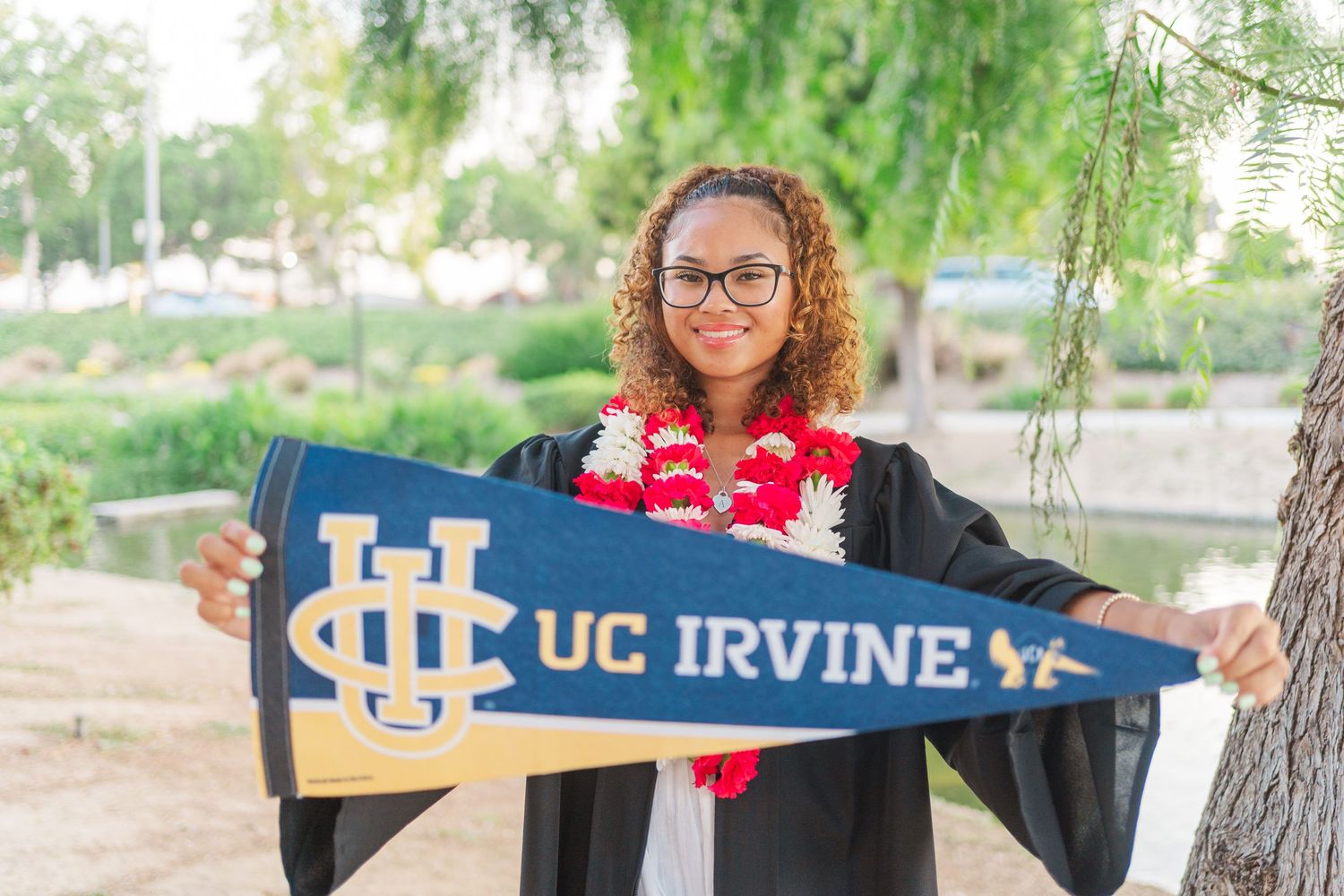 Graduate in cap and gown displays UC Irvine pennant with red lei while standing under a willow tree.