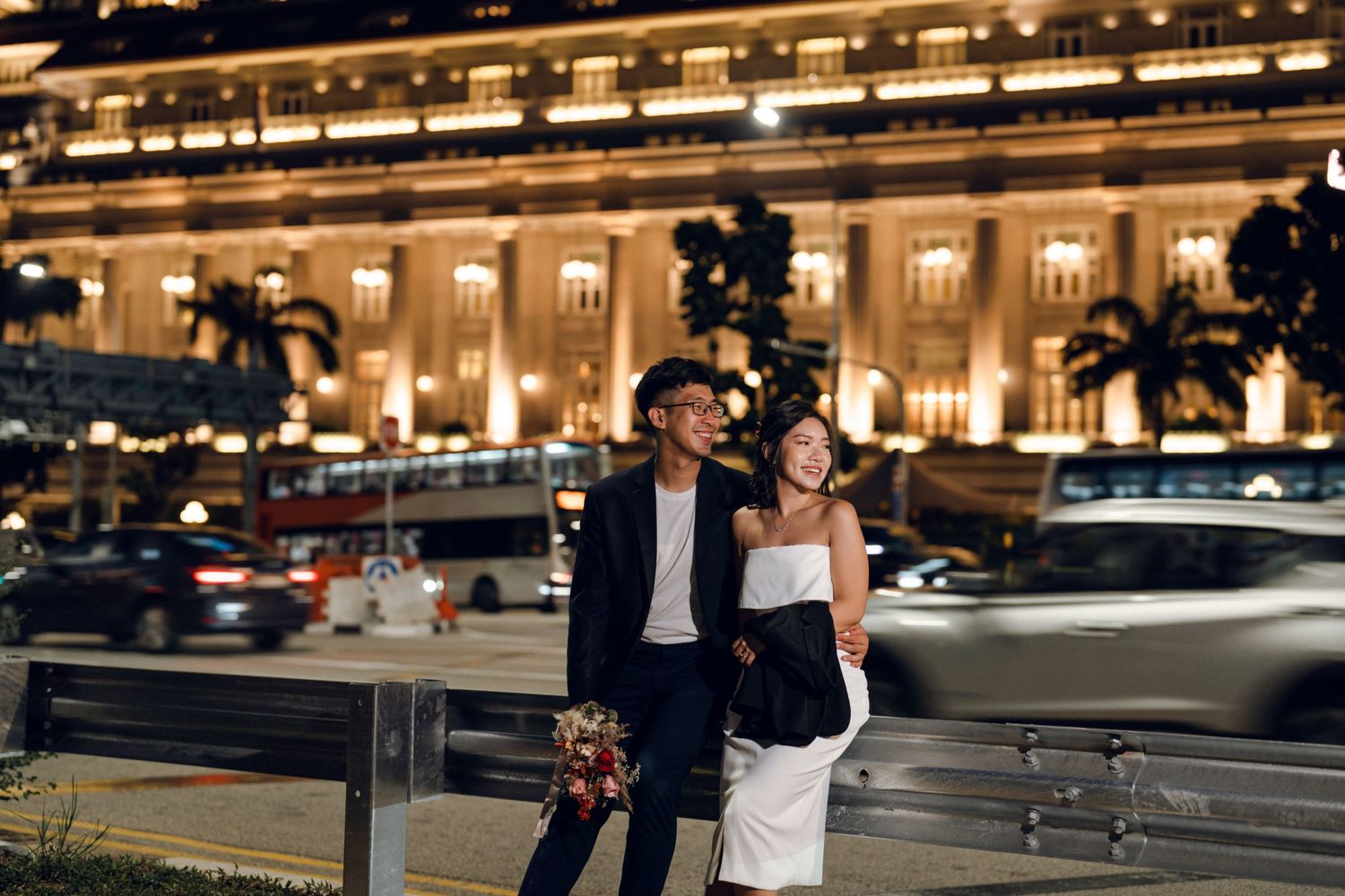 Romantic nighttime photoshoot in front of the illuminated Fullerton Hotel with city lights in the background.