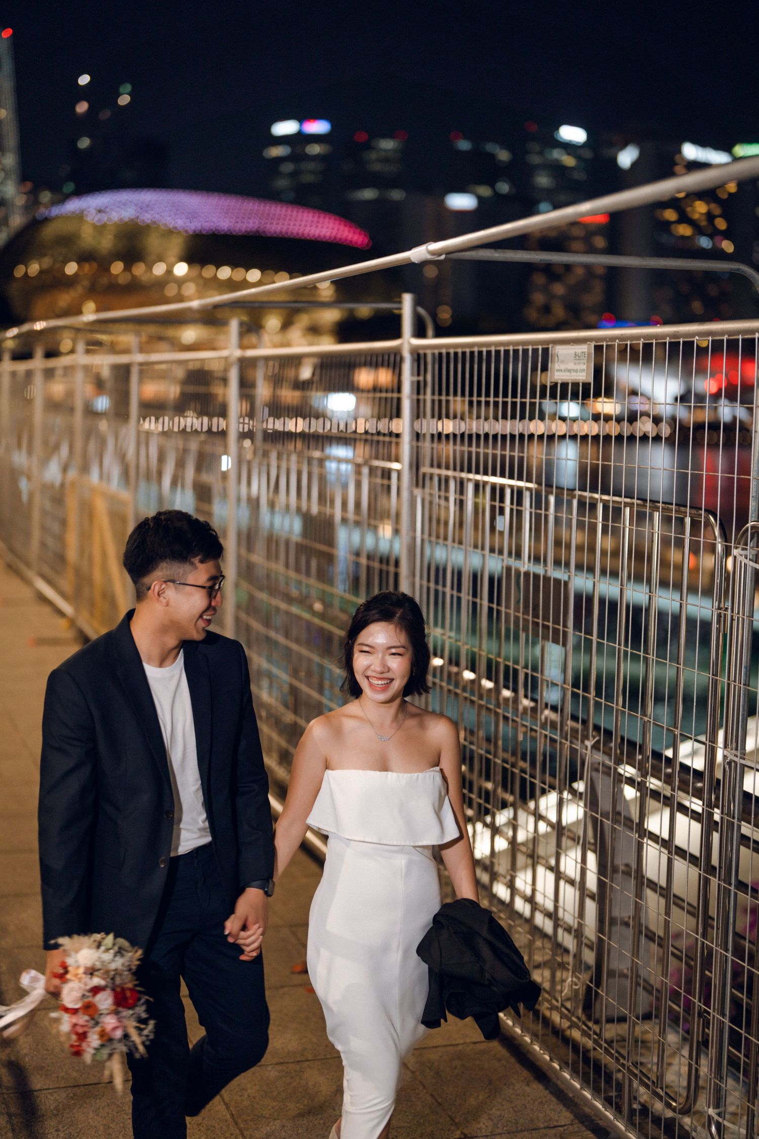 Couple walking and laughing along illuminated walkway with metal railings at night.