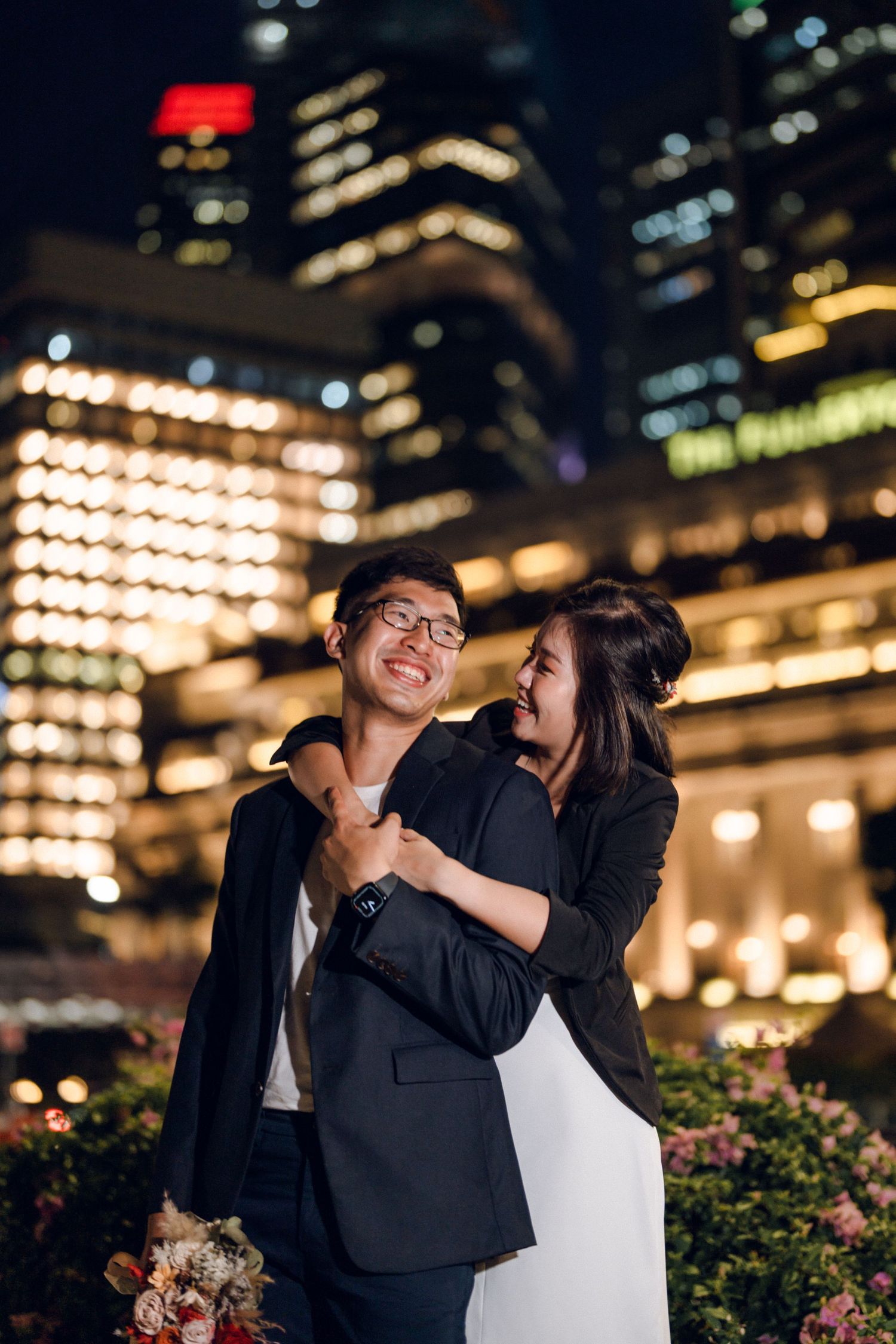 Joyful couple embracing against nighttime cityscape with decorative lighting.