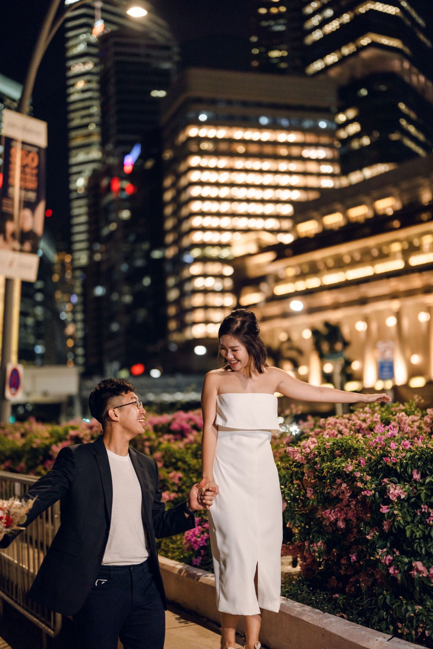 Romantic couple poses against urban nightscape with blooming pink flowers in foreground.