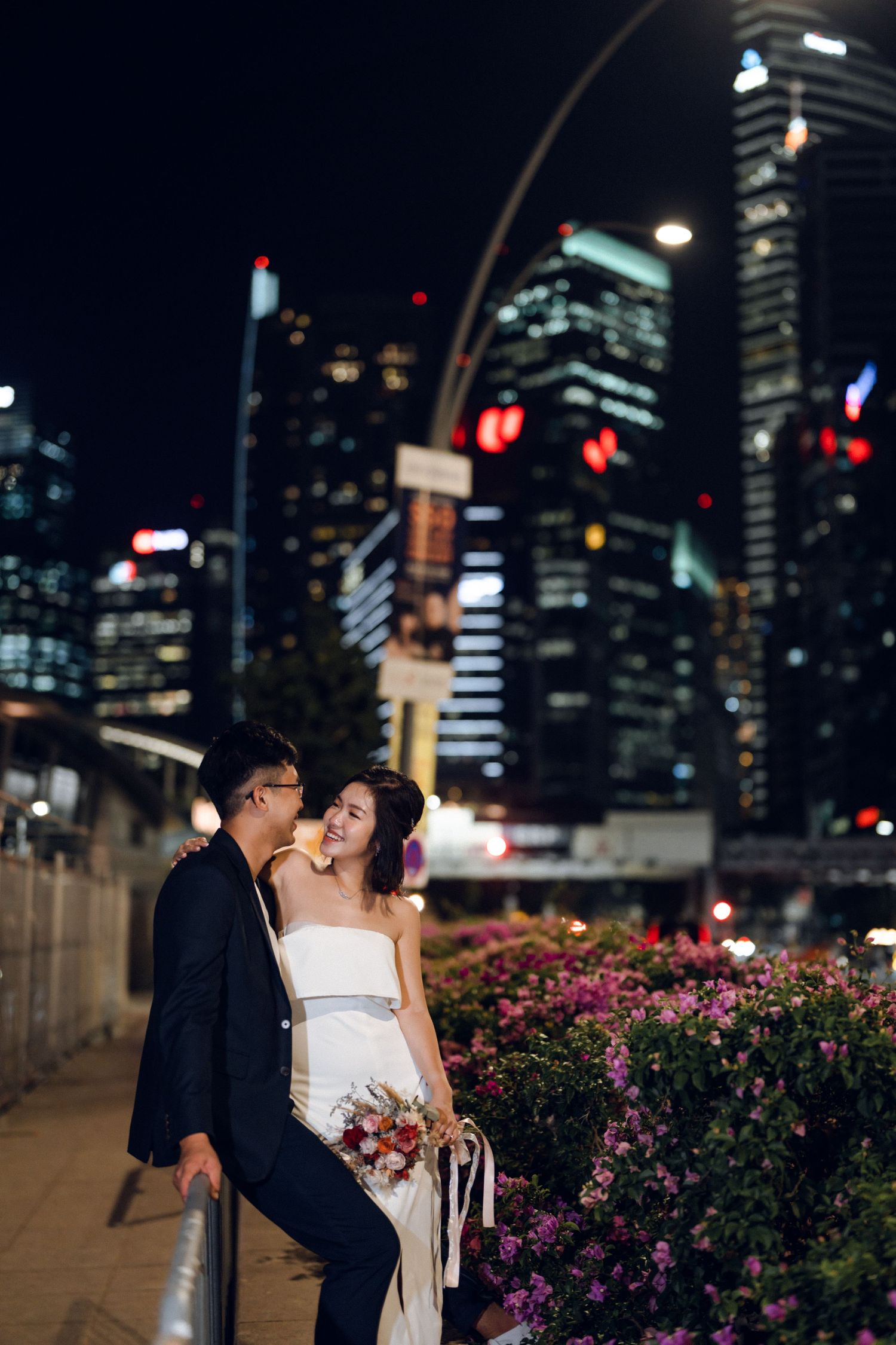 Romantic nighttime portrait with city lights and flowering bushes creating dreamy atmosphere.