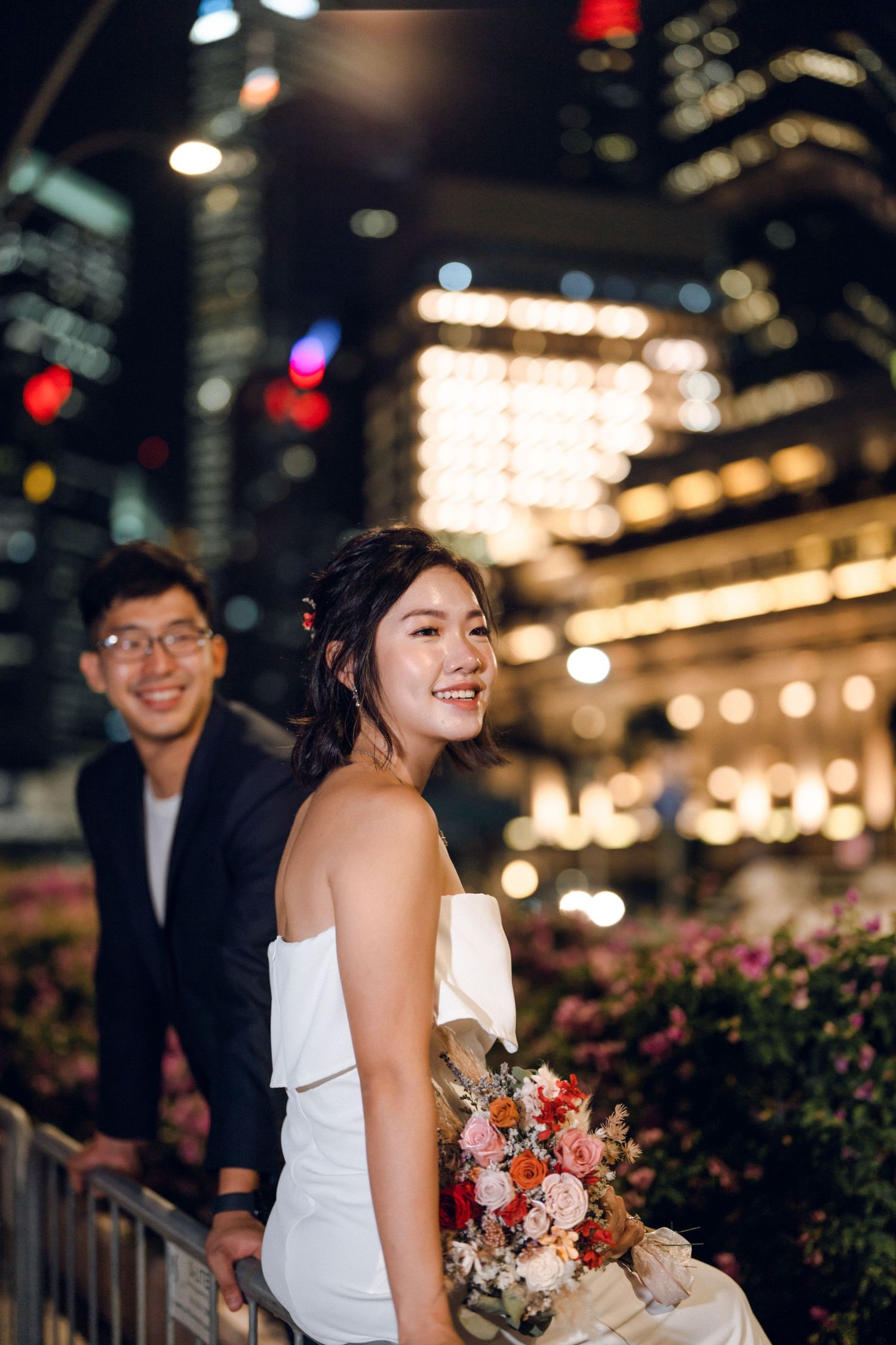 Joyful evening photoshoot with bouquet against illuminated urban backdrop.