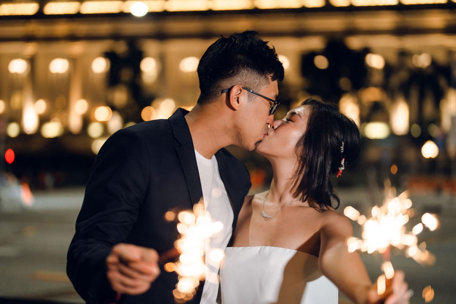 Couple embracing during a romantic sparkler-lit evening with city lights in background.