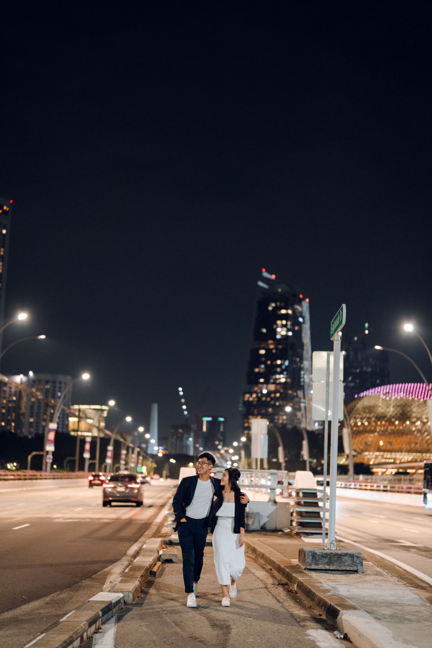 Couple walking along illuminated city street at night with skyline in background.