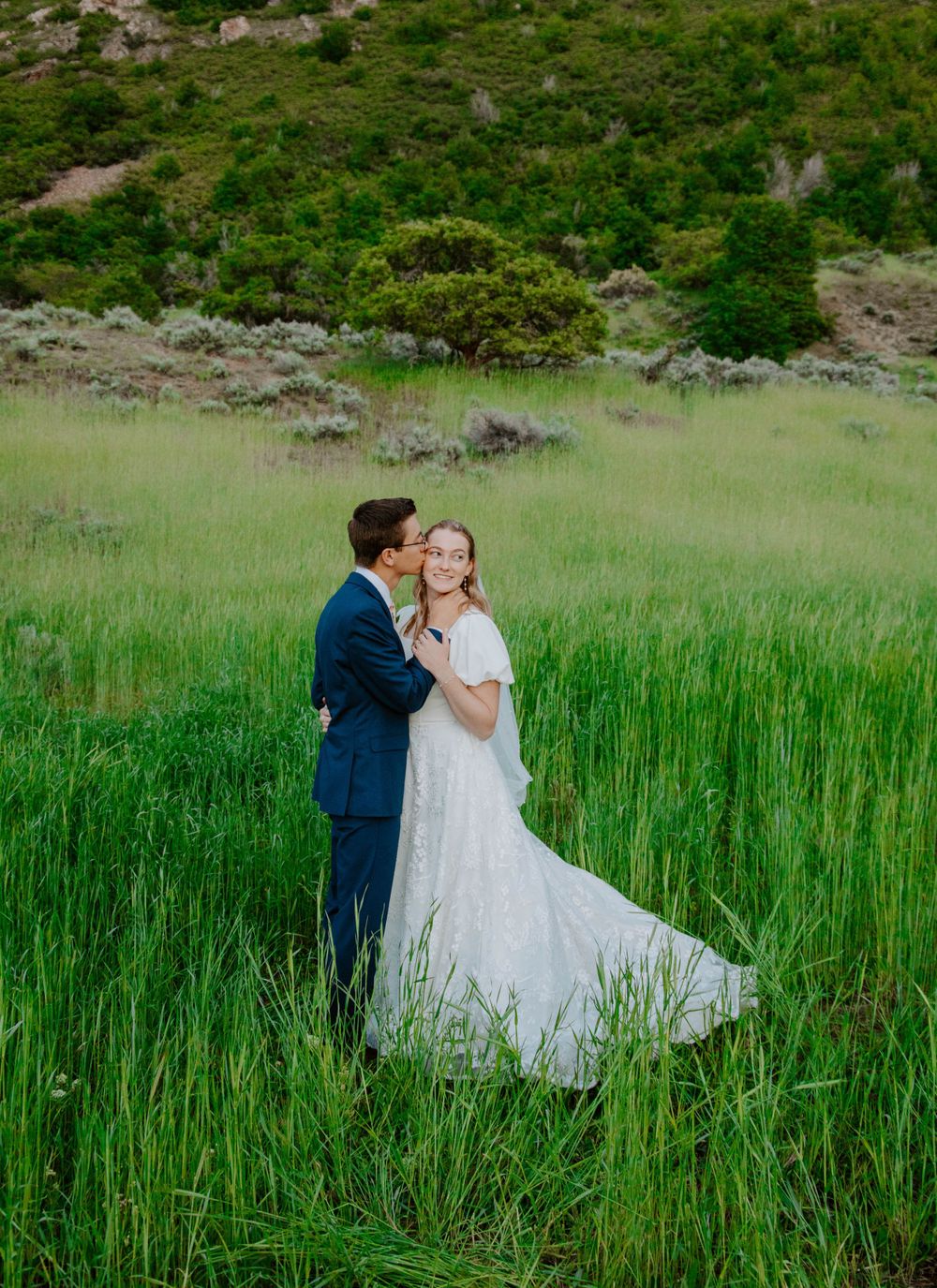 Couple shares intimate moment in tall green grass with mountains in background.
