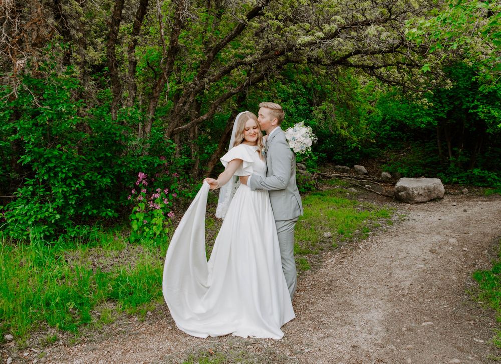A couple in wedding attire embraces on a nature path surrounded by spring greenery.