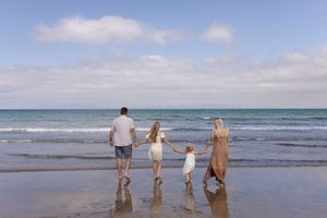 Family walking along sandy beach shoreline with waves and cloudy sky in the background.