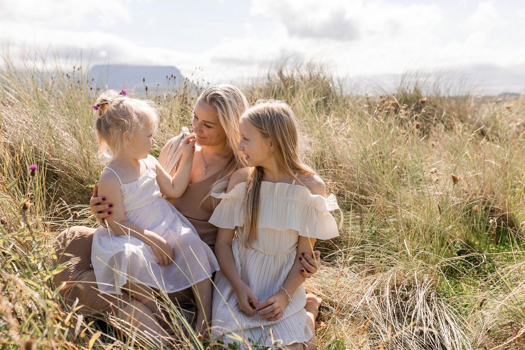 A group in white outfits sits together in a sunlit meadow with mountains visible in the distance.