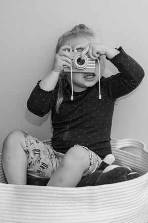 A young child in polka dot shirt holds up a toy camera while sitting in a white woven basket, captured in black and white.