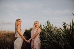 Two people in light colored dresses holding hands at sunset near tropical plants.
