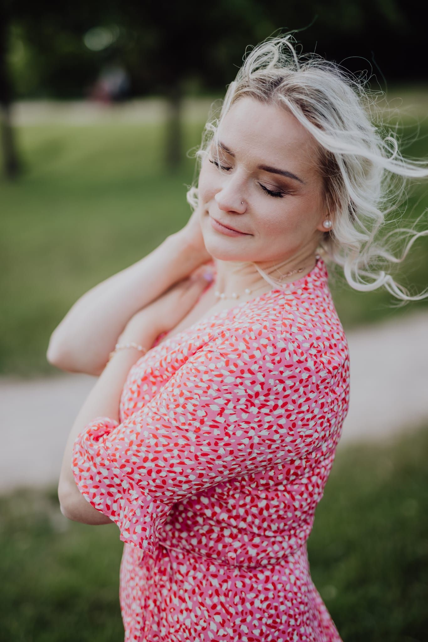 Woman in pink floral dress with windswept blonde hair posing outdoors.