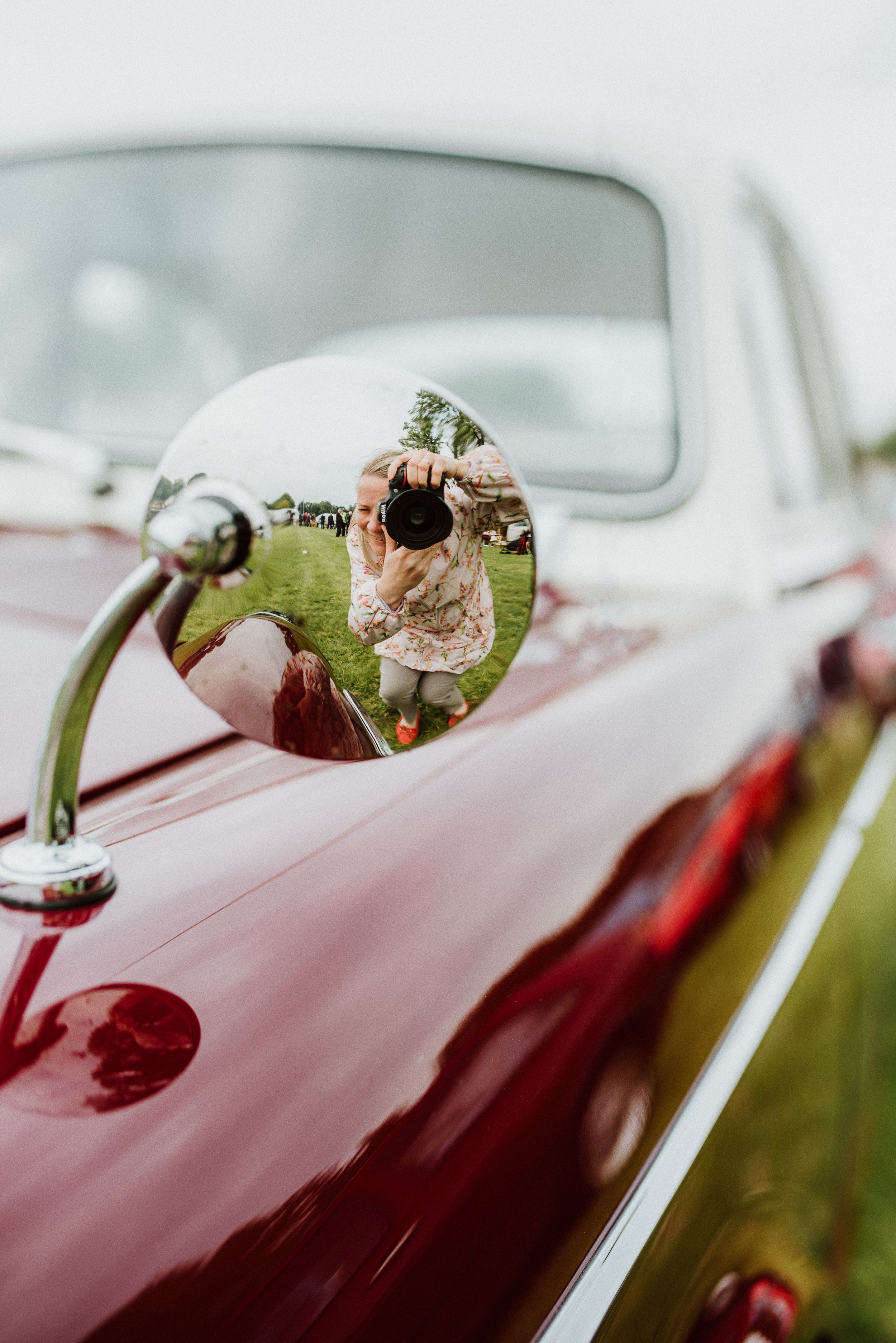 Reflection in vintage car side mirror shows person in light dress on grassy field. Self-portrait. Artsy.
