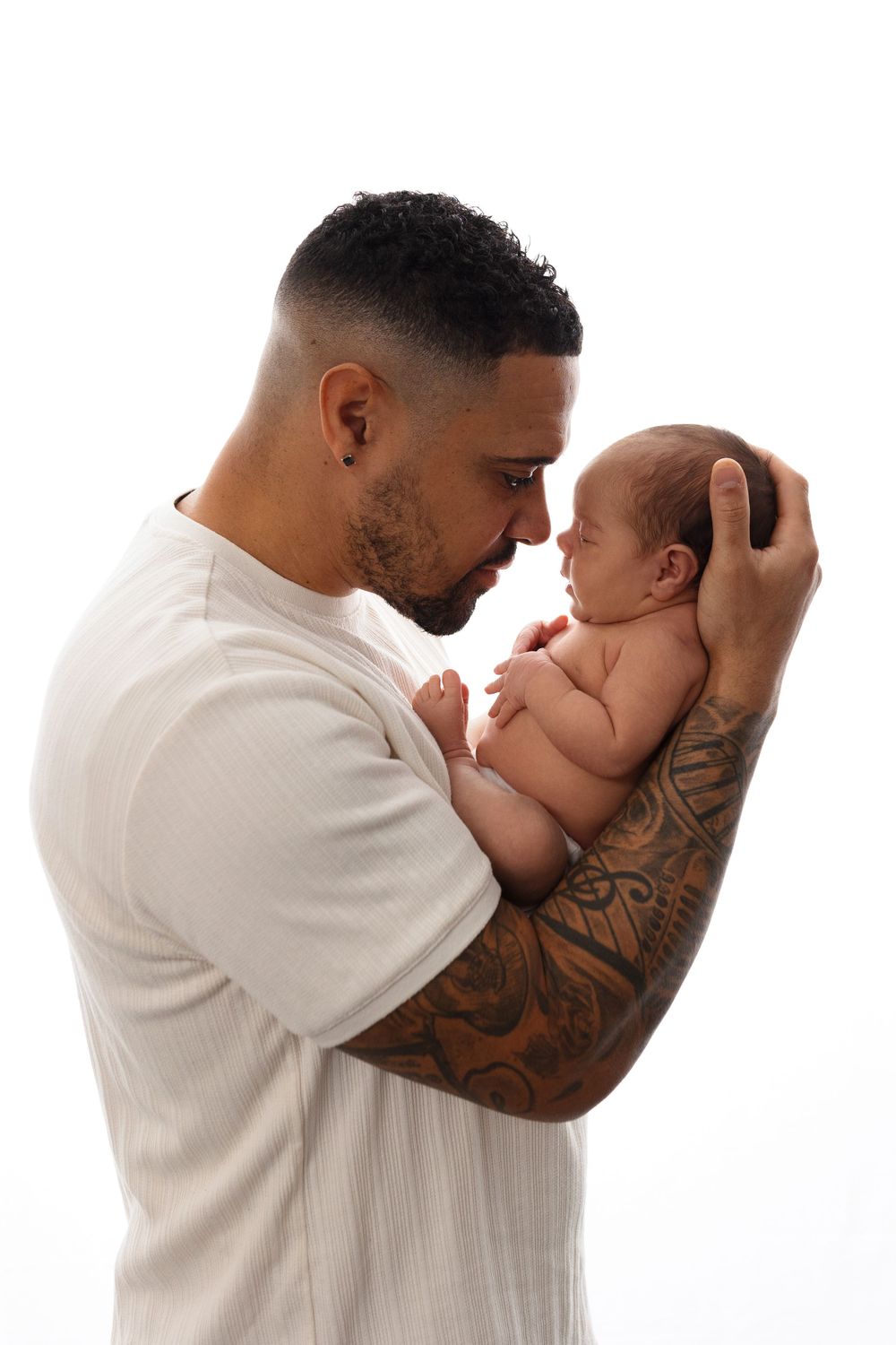 Tattooed father in white shirt tenderly holding newborn baby against white background.