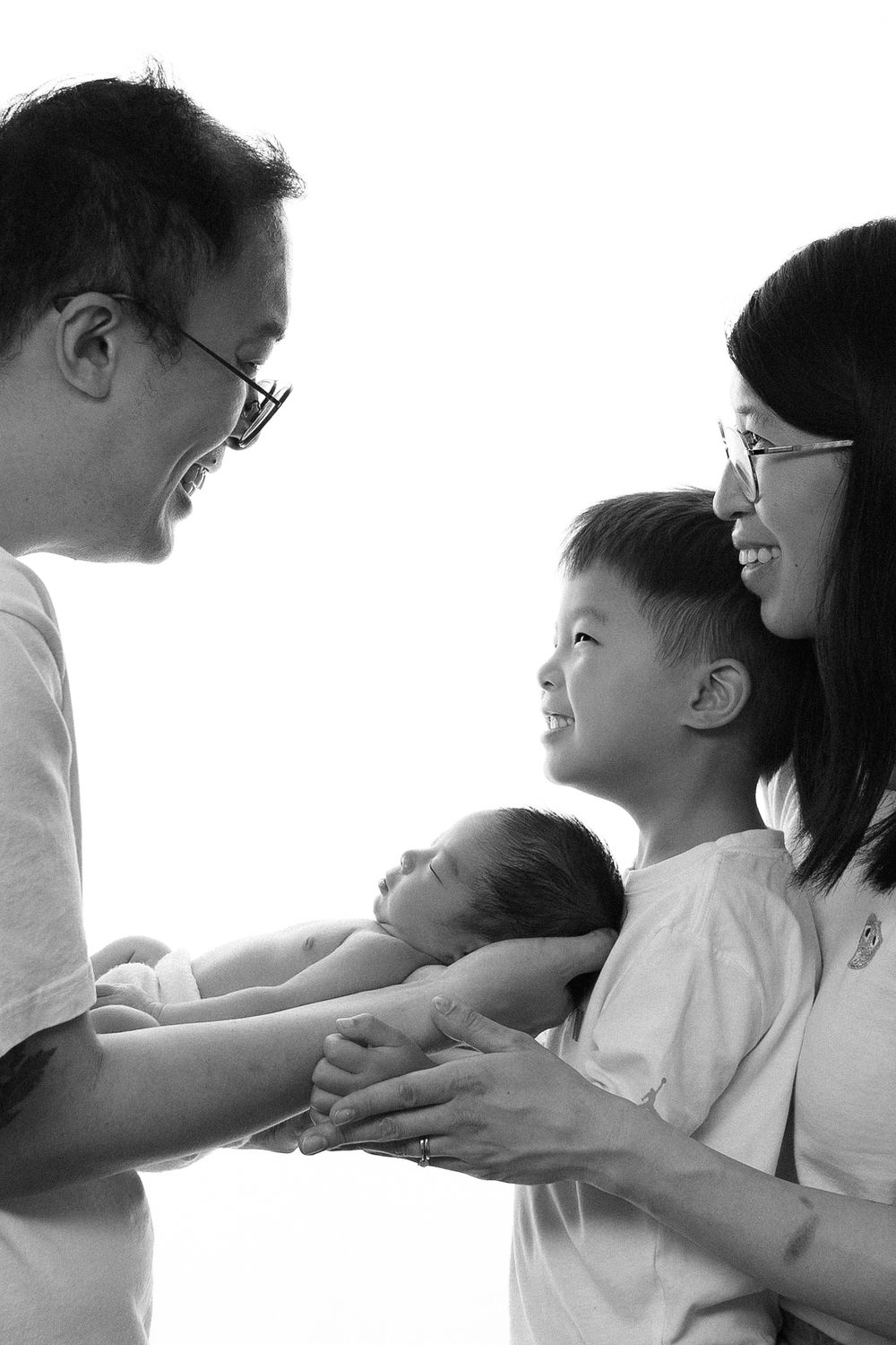 Black and white family portrait with parents smiling at young boy holding newborn baby.