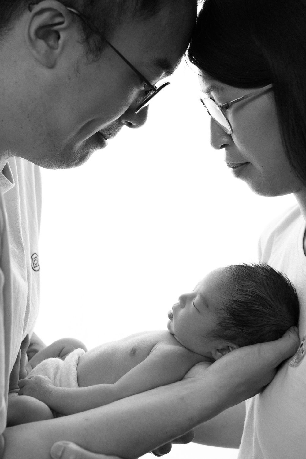 Black and white photo of two parents wearing glasses lovingly gazing at their sleeping newborn baby.