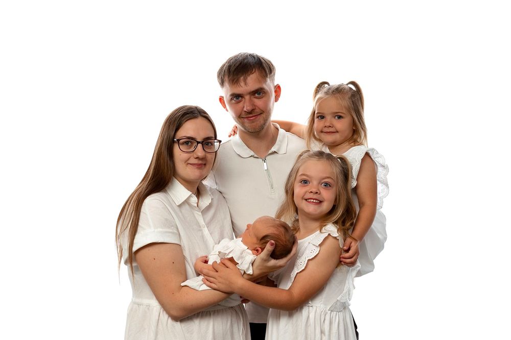 A happy family of four in white outfits poses together, holding a newborn baby against a white background.
