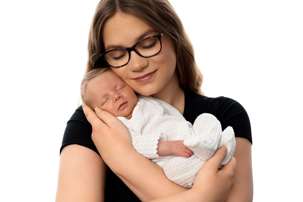 A parent in glasses tenderly holds and gazes at their sleeping newborn baby wrapped in white.