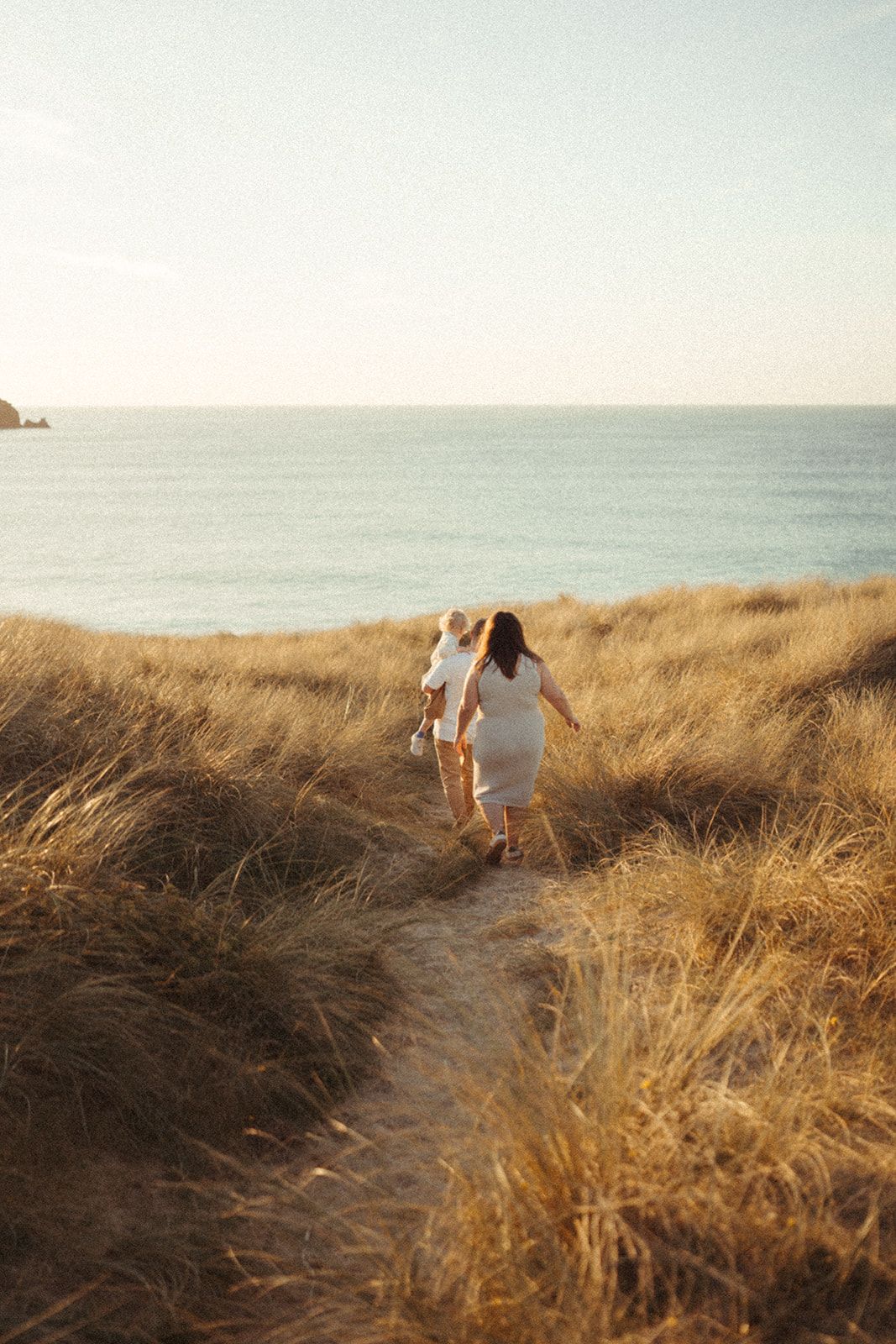 Two people walking up a coastal trail through golden grass at sunset with the ocean horizon in view.