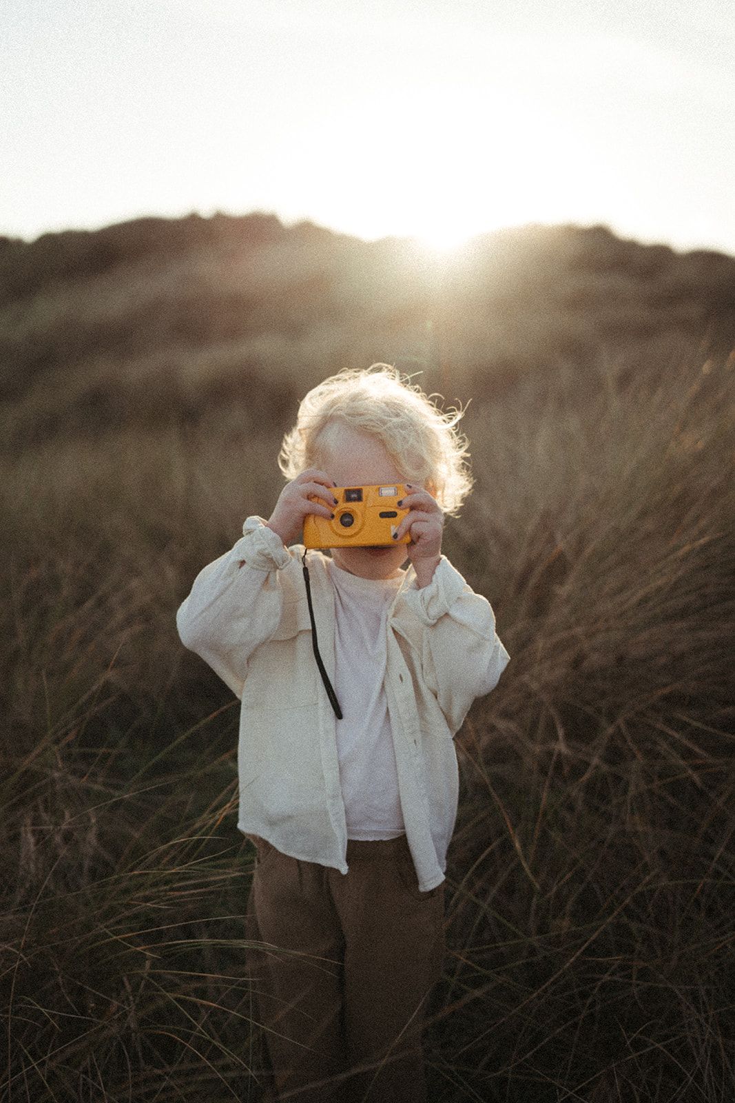 Child holds yellow toy camera standing in tall grass at golden hour with sun setting behind.