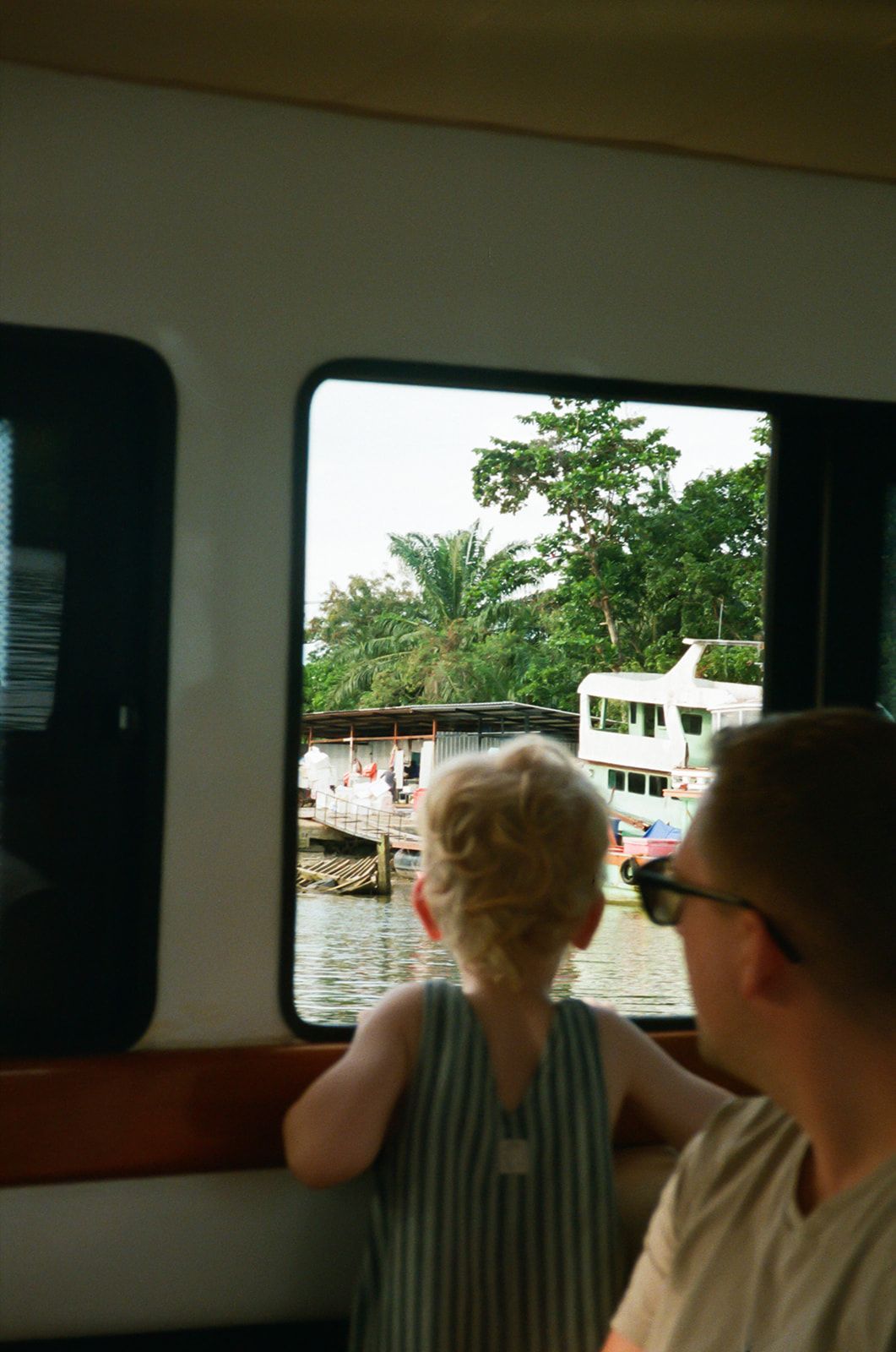 Two passengers look out through a boat window at docked vessels and tropical greenery along a waterfront.
