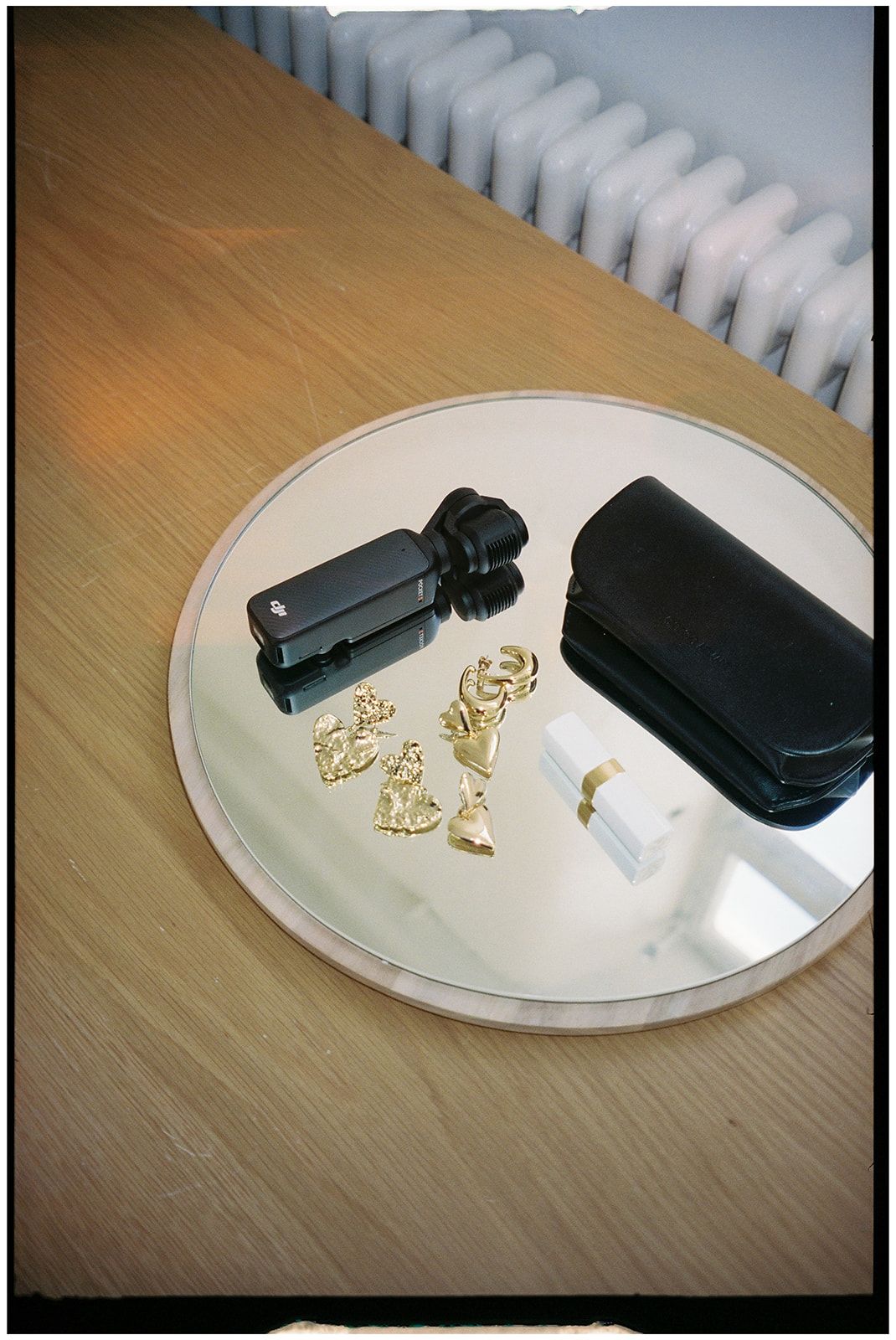 A black camera and case arranged on a round mirror surface with scattered gold jewelry pieces on wooden table.