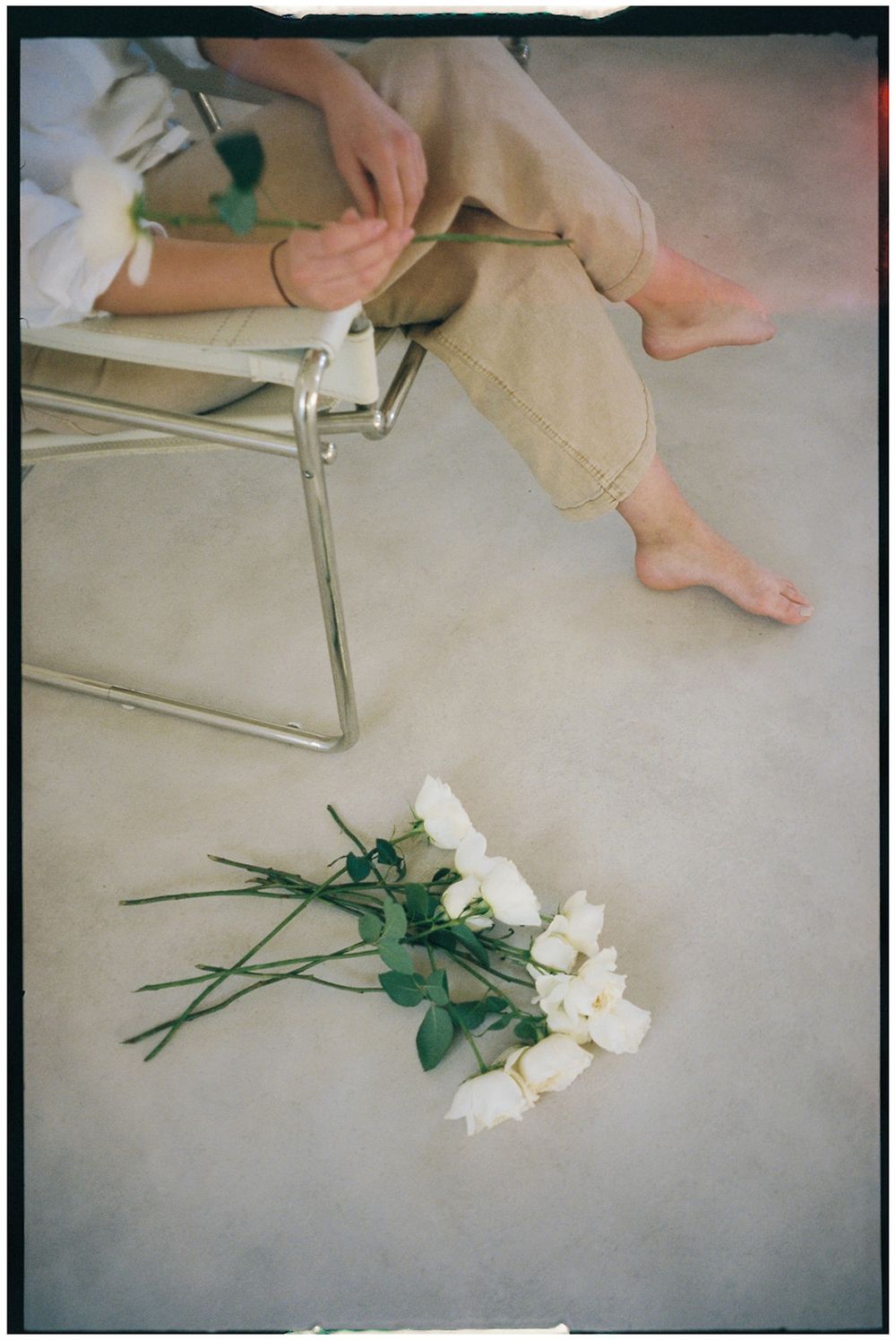 Artistic photograph of legs resting on a metal chair with white roses and greenery on beige carpet.