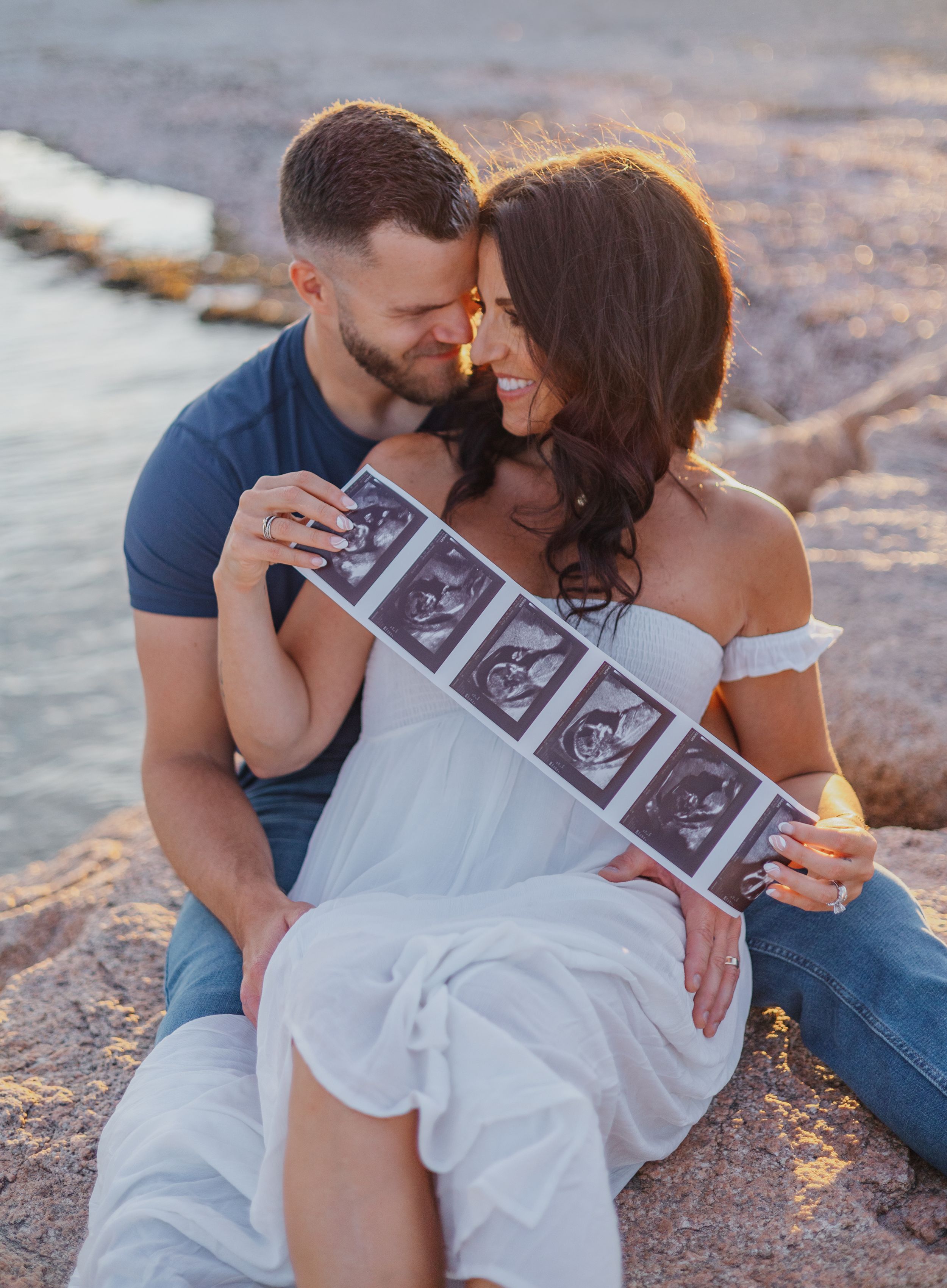 Couple sharing intimate moment while holding ultrasound photos at sunset.