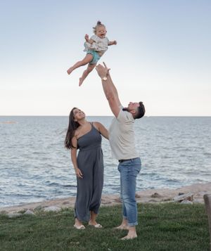 Parents lifting child in the air playfully by ocean at sunset.