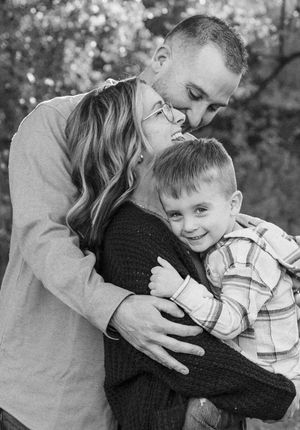 Sweet family moment outdoors with adults embracing while young boy in plaid shirt smiles at camera in black and white.