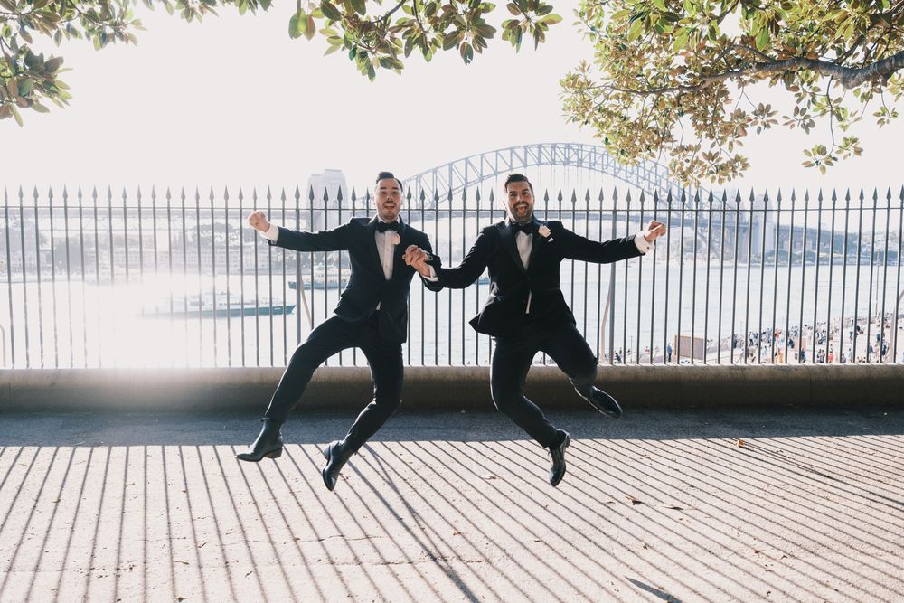 Two grooms in tuxedos joyfully jumping in front of Sydney Harbour Bridge on their wedding day.