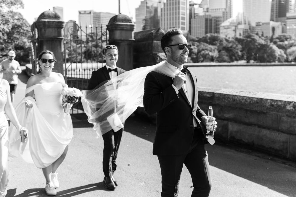 Black and white wedding photo of groom holding veil in wind, walking with bridal party near waterfront with city skyline.