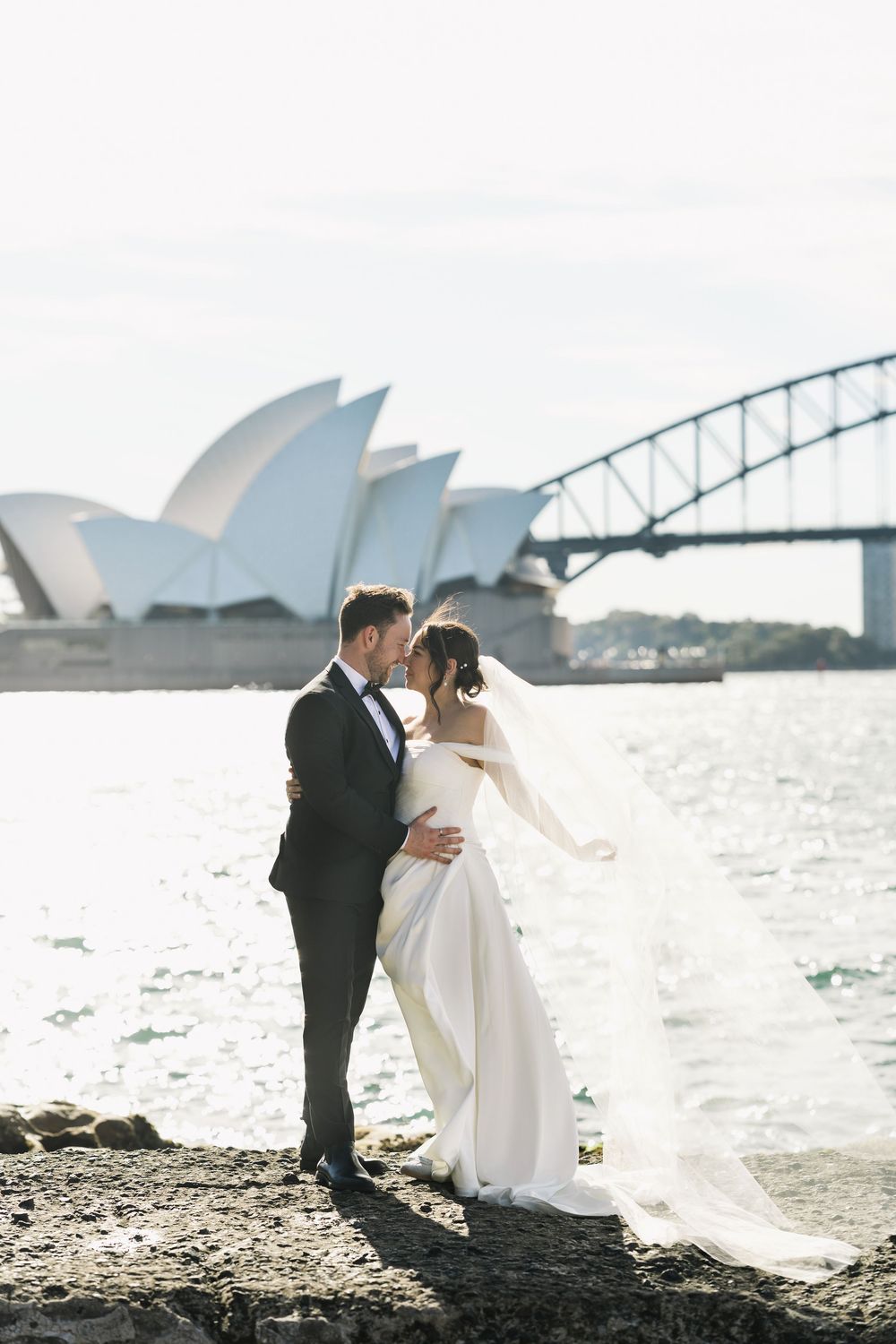 Bride and groom share a romantic moment by Sydney Harbour with the Opera House and Harbour Bridge in the background.