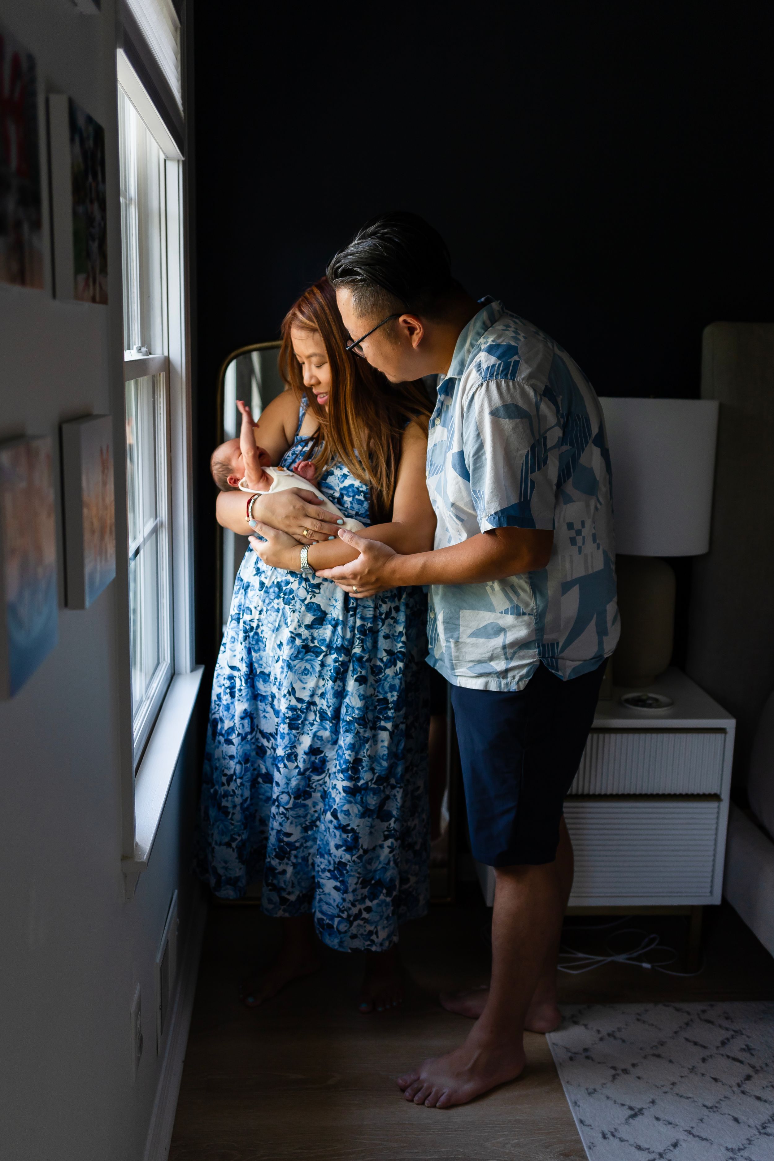 Family standing by window sharing intimate moment with newborn baby in blue floral dress.