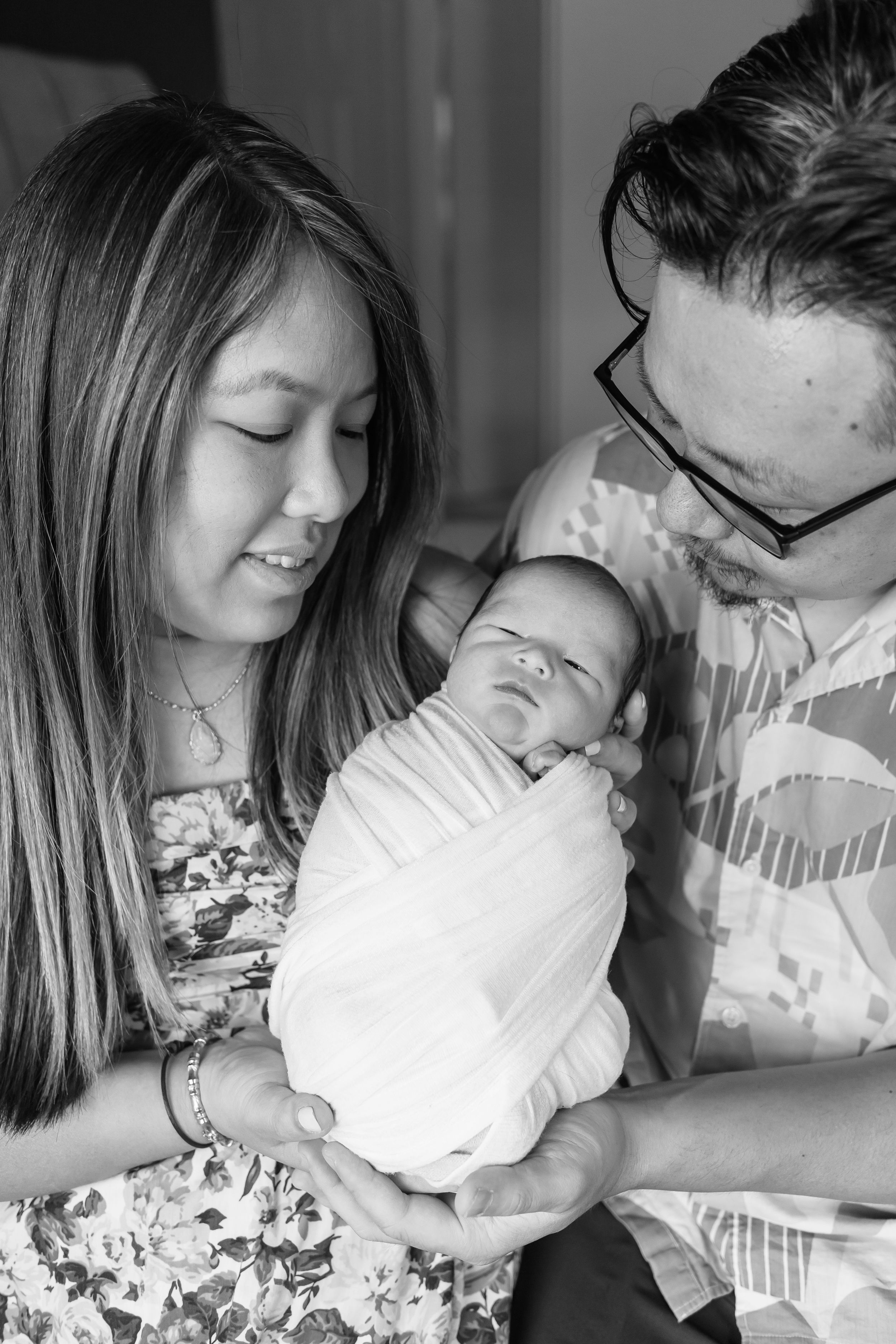 Black and white portrait of family holding sleeping newborn wrapped in white.
