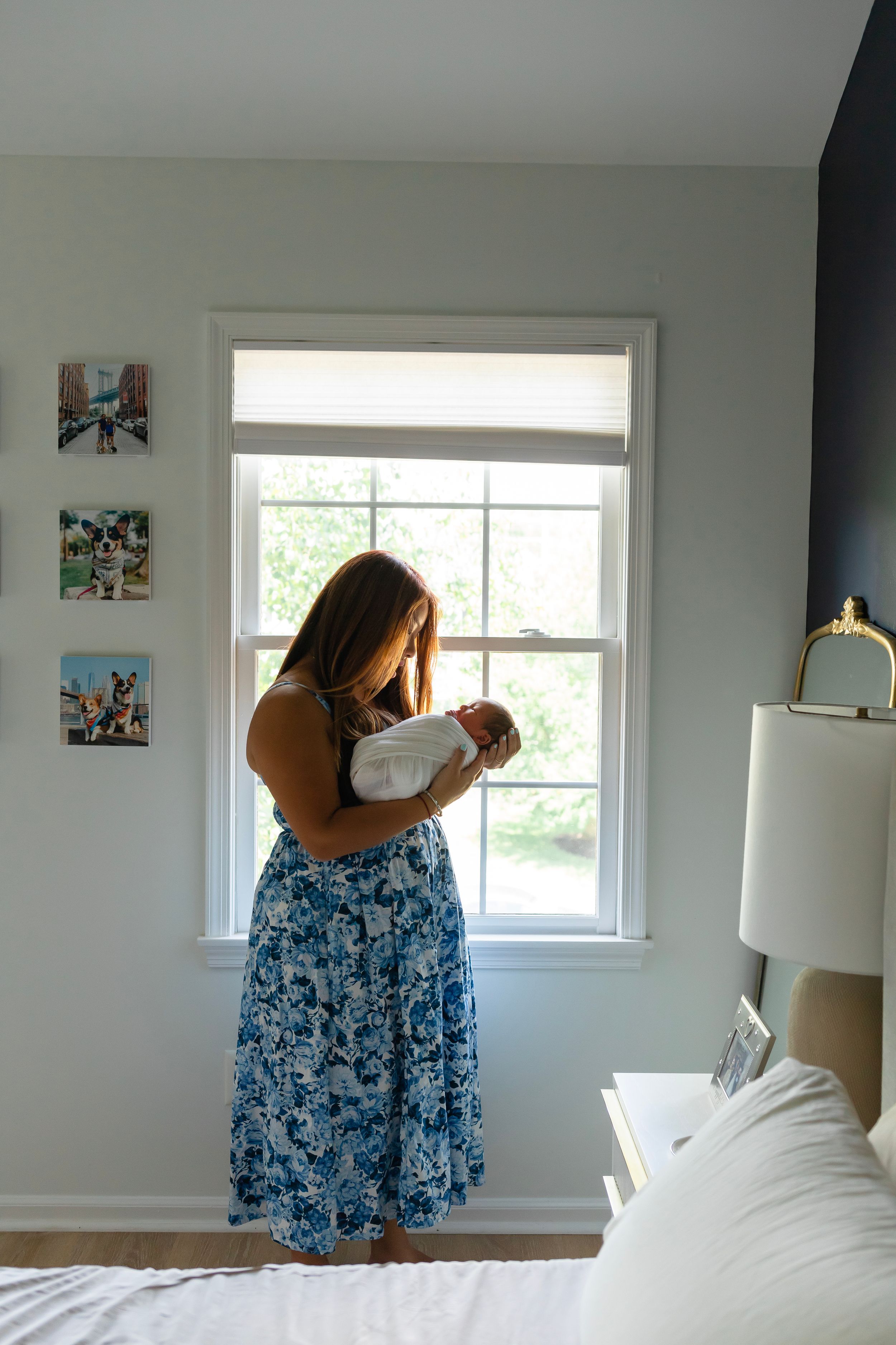 Person in floral dress holds newborn baby while standing by bright window.