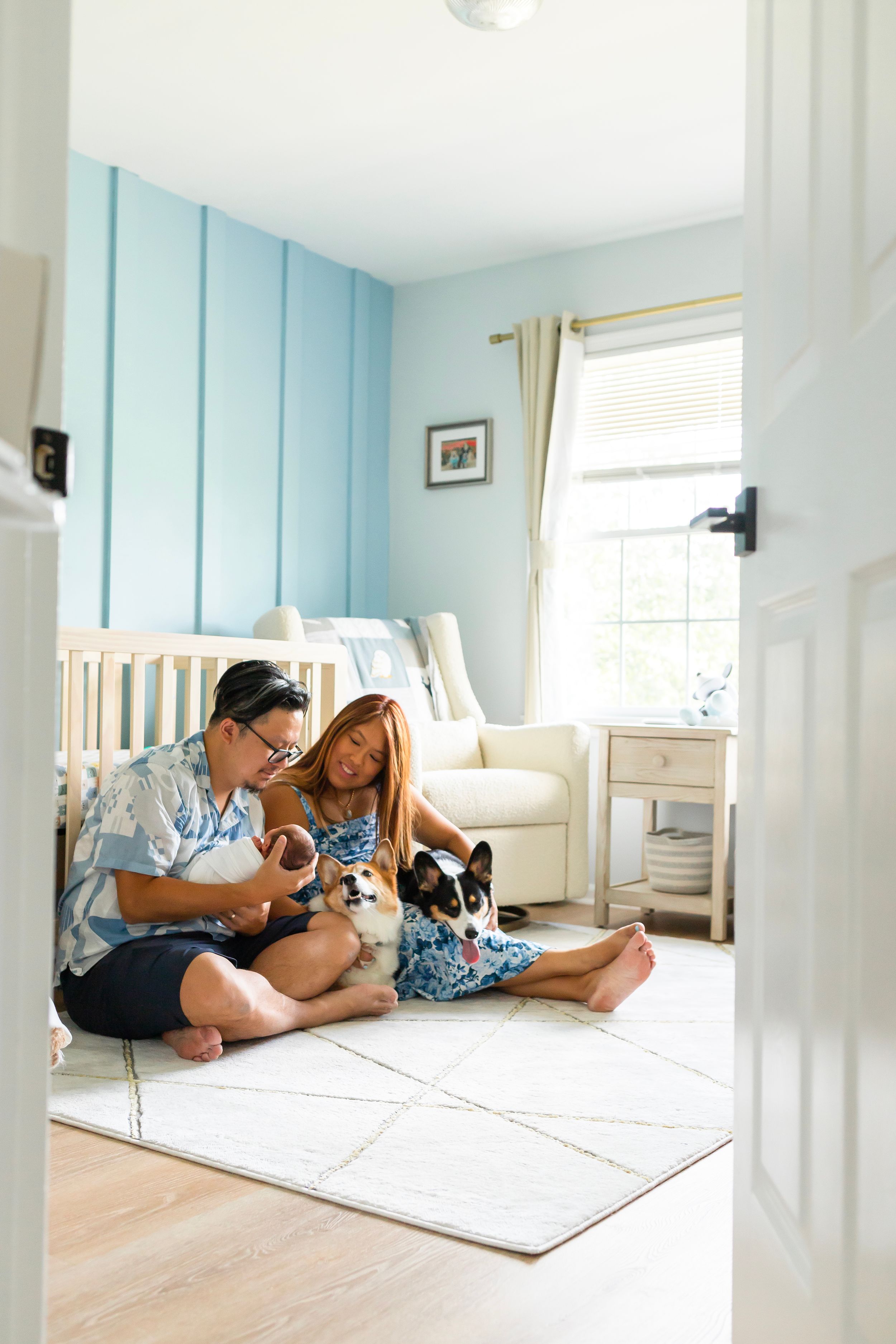 A couple sits on a nursery floor with their dogs while holding a baby.