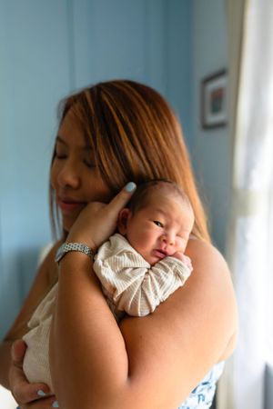 A swaddled newborn baby rests peacefully on a shoulder in natural window light.
